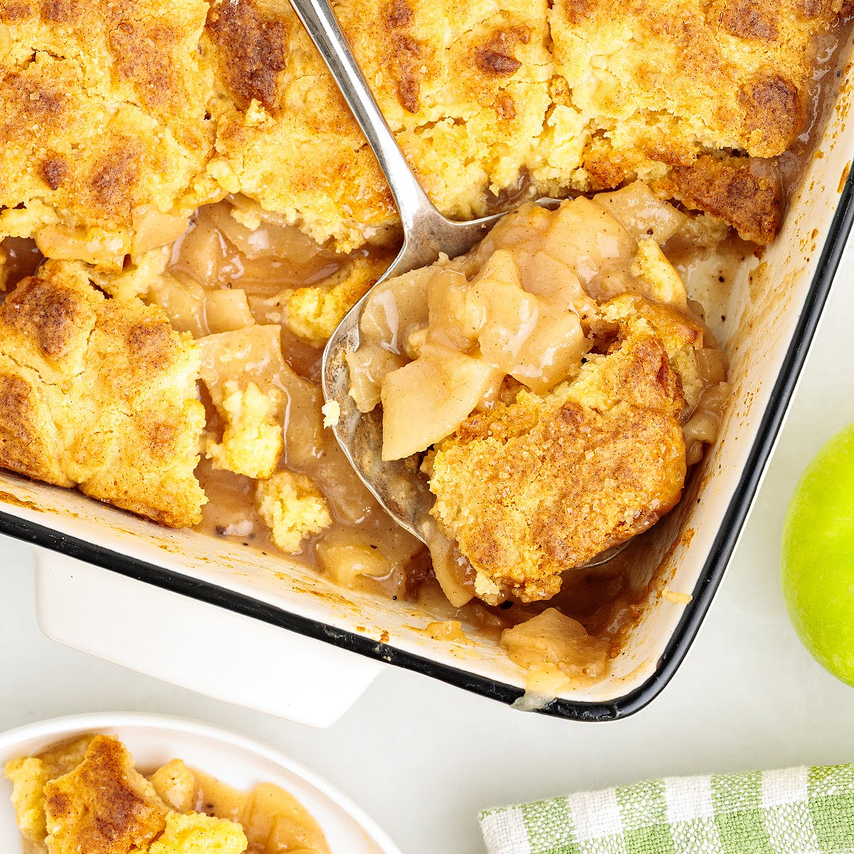 Apple cake cobbler with cinnamon-spiced Granny Smith apple filling and golden butter cake topping being served from the baking dish.
