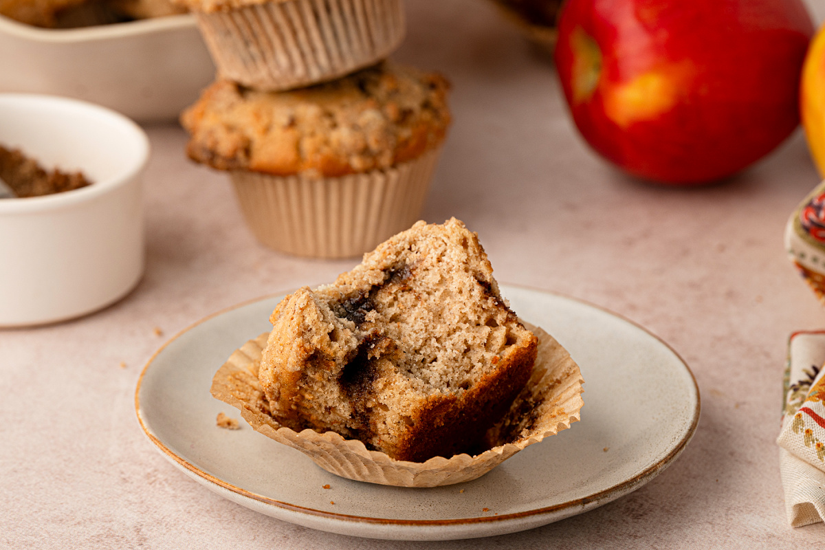 Apple cider streusel muffins made with reduced fresh cider, warm spices, brown sugar cinnamon and buttery streusel. Apple cider streusel muffin broken open on a plate with whole apples and more muffins in the background.