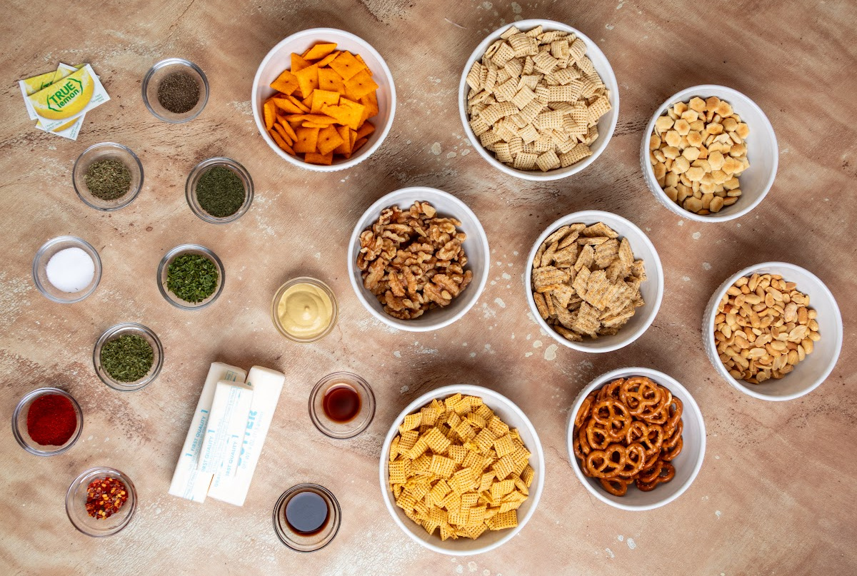 Ingredients for cowboy butter snack mix including Chex cereal, Cheez-It crackers, Triscuits, pretzels, peanuts, walnuts, oyster crackers, butter, herbs and seasonings