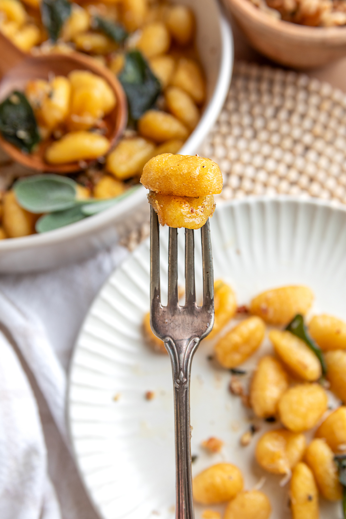 Close-up of the browned exterior on crispy potato gnocchi. Crispy gnocchi with sage brown butter on a fork with more pan-fried gnocchi and sage in the background.