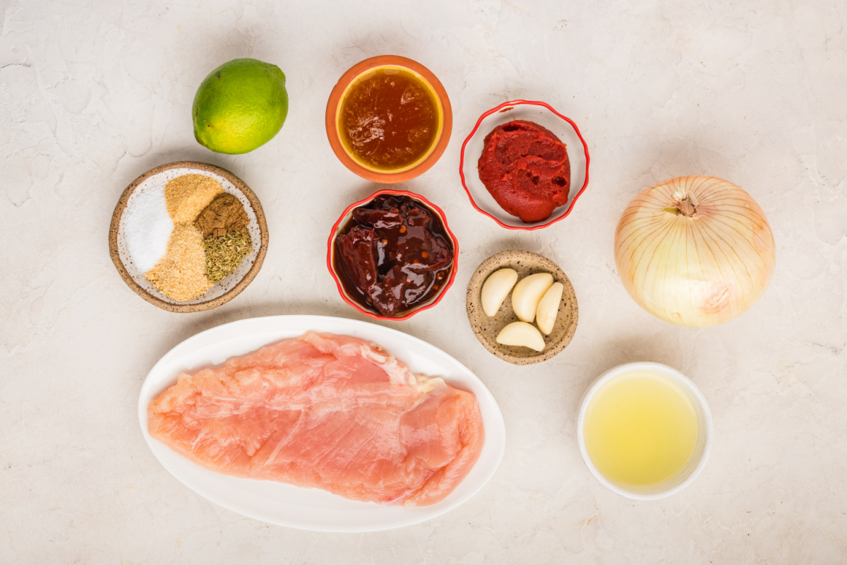 Ingredients for slow cooker chipotle chicken including chicken breasts, chipotle peppers, tomato paste, lime, onion and seasonings