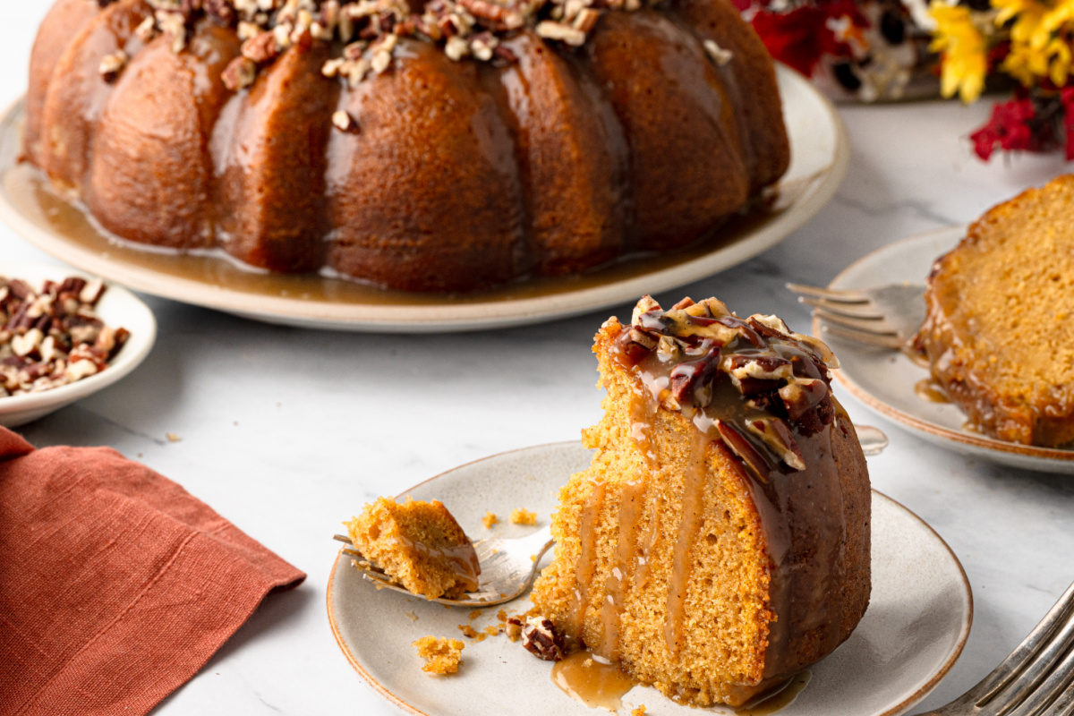 Sweet potato Bundt cake made with roasted sweet potatoes and brown butter, served with rum caramel and toasted pecans. Slice of sweet potato Bundt cake on a plate with buttered rum caramel dripping down the cut side and toasted pecans on top.