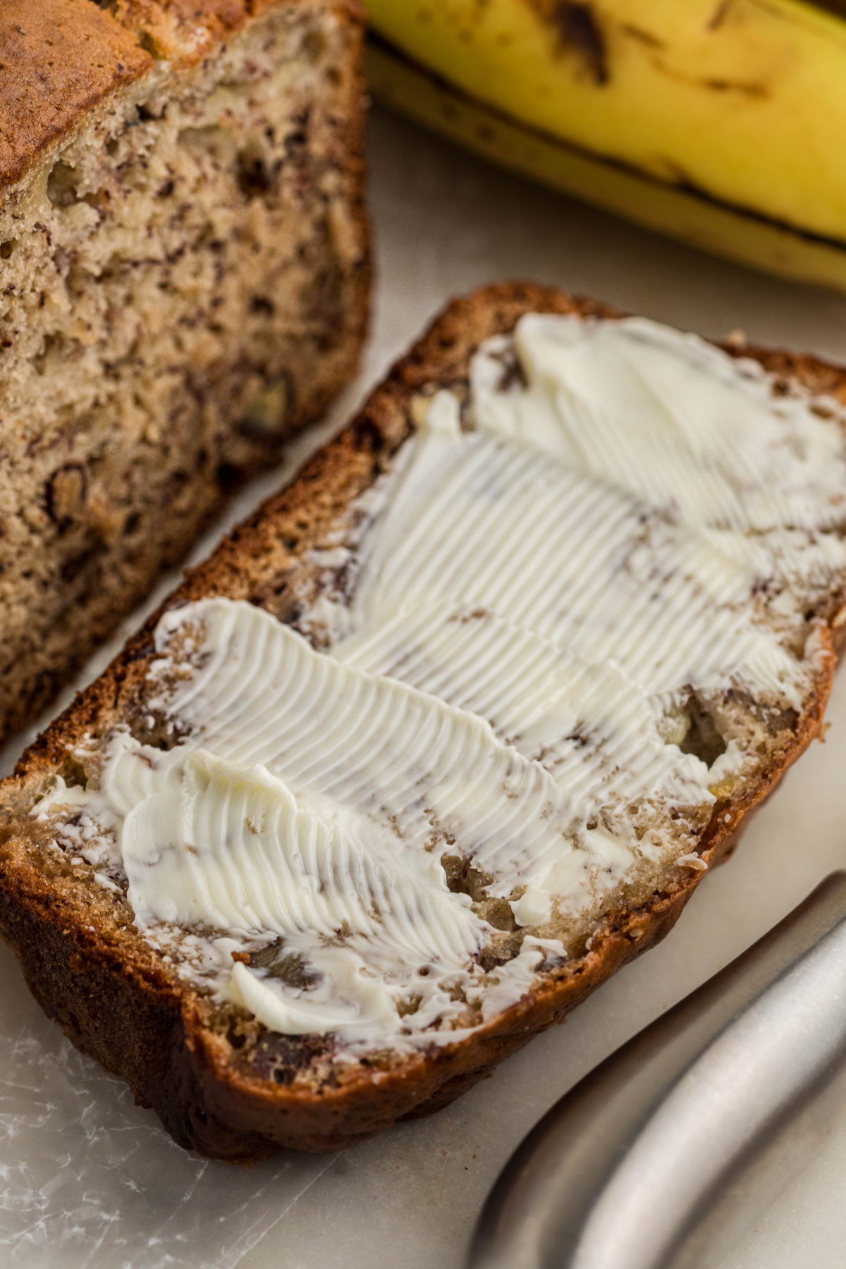 Thick slice of banana bread with cottage cheese spread with butter, with ripe bananas in the background.