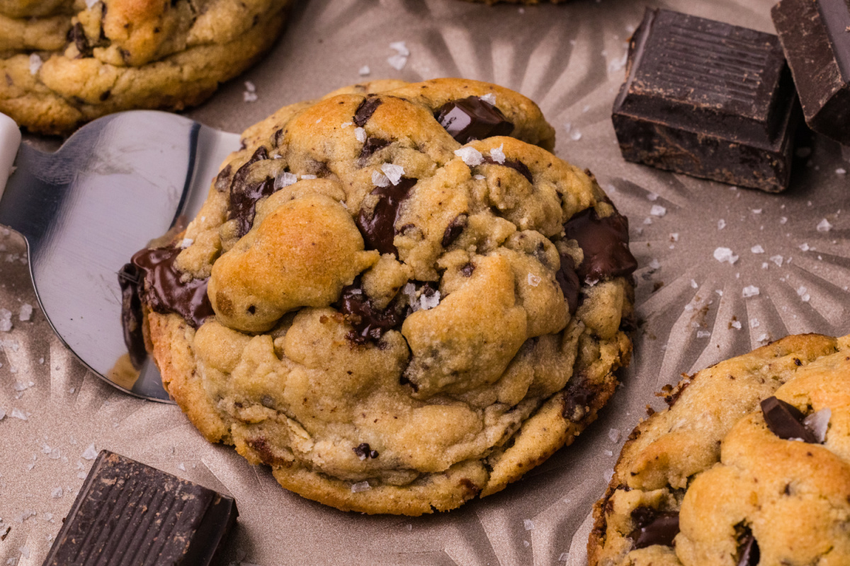 Thick chocolate chip cookie resting on a metal spatula with melted dark chocolate, golden dough, and flaky sea salt.