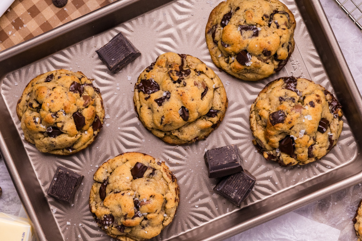 Five thick brown butter chocolate chip cookies with golden tops, dark chocolate chunks, and flaky sea salt on a metal baking sheet.