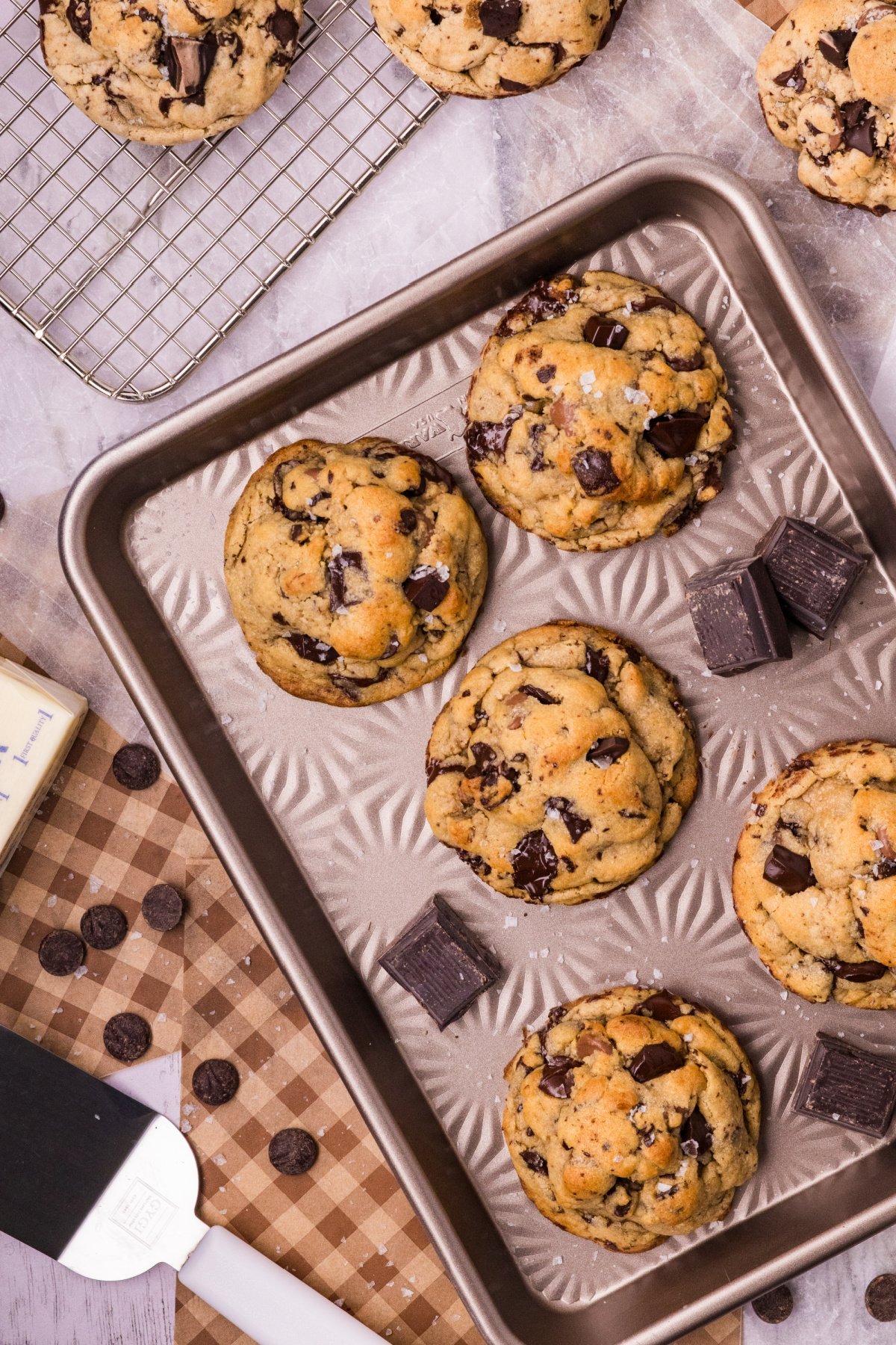Thick chocolate chip cookies with golden tops, dark chocolate chunks, and flaky sea salt arranged on a baking sheet and wire cooling rack.