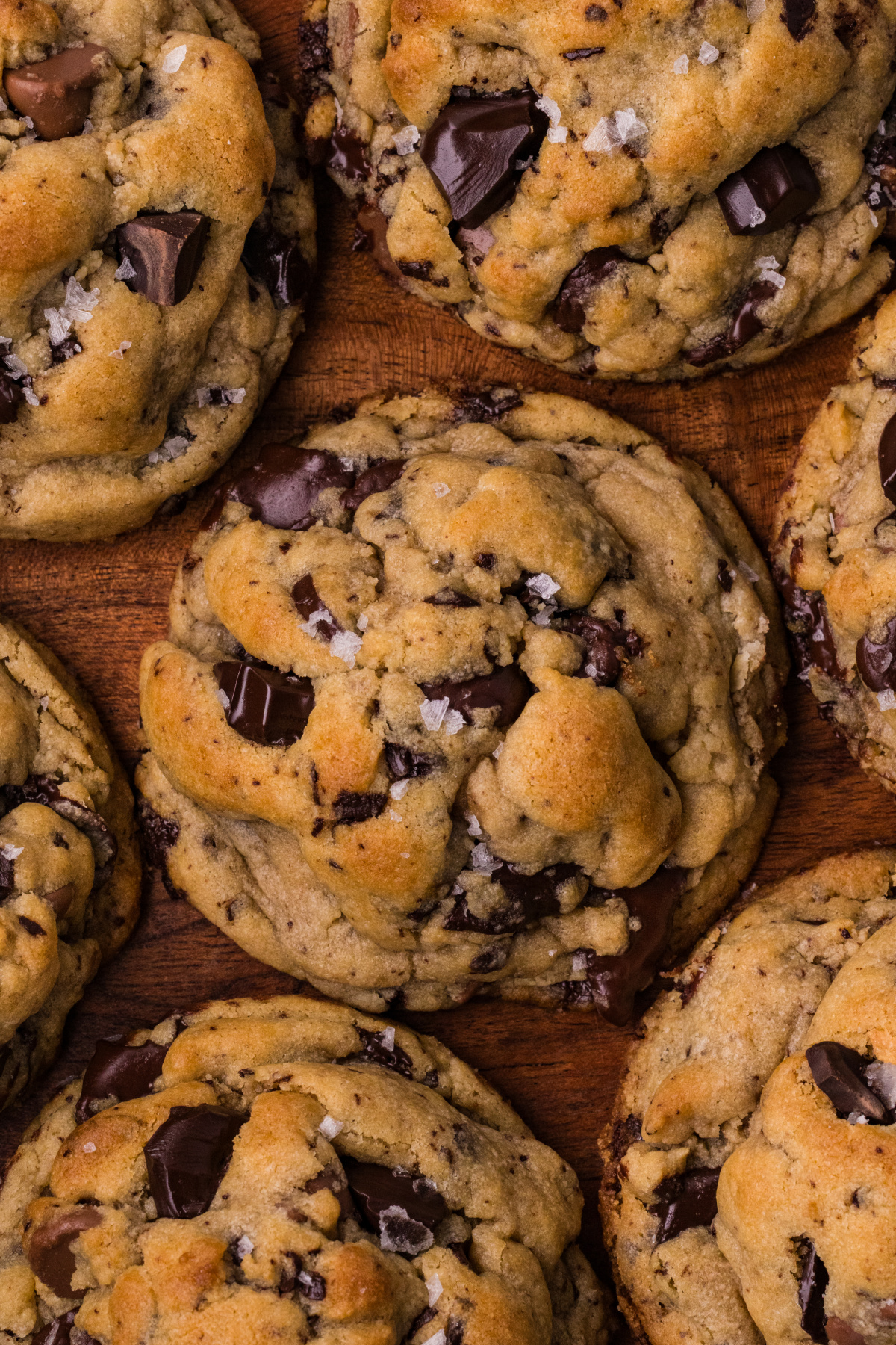 Brown butter chocolate chip cookies with golden tops, melted dark and milk chocolate, and flaky sea salt on a wooden board.