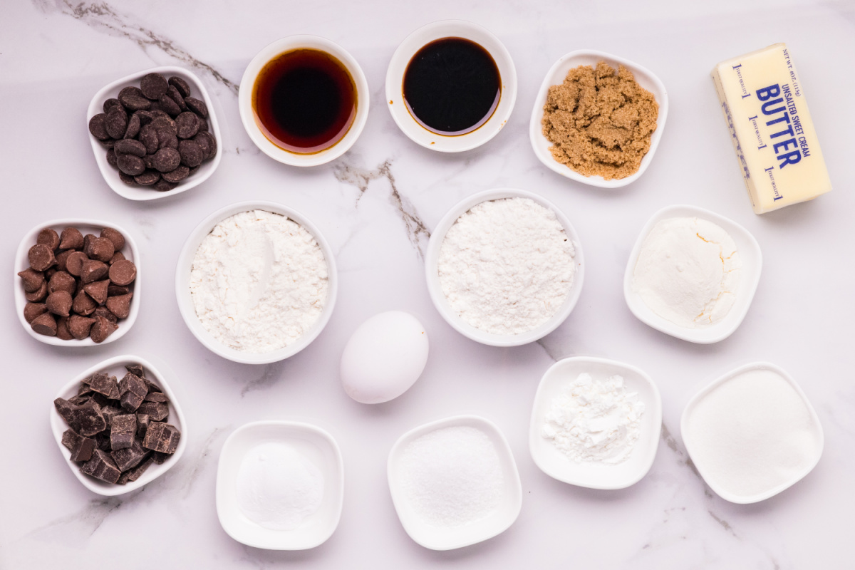 Ingredients for brown butter chocolate chip cookies arranged in bowls, including butter, two flours, sugars, dry milk powder, molasses, egg, and three kinds of chocolate.