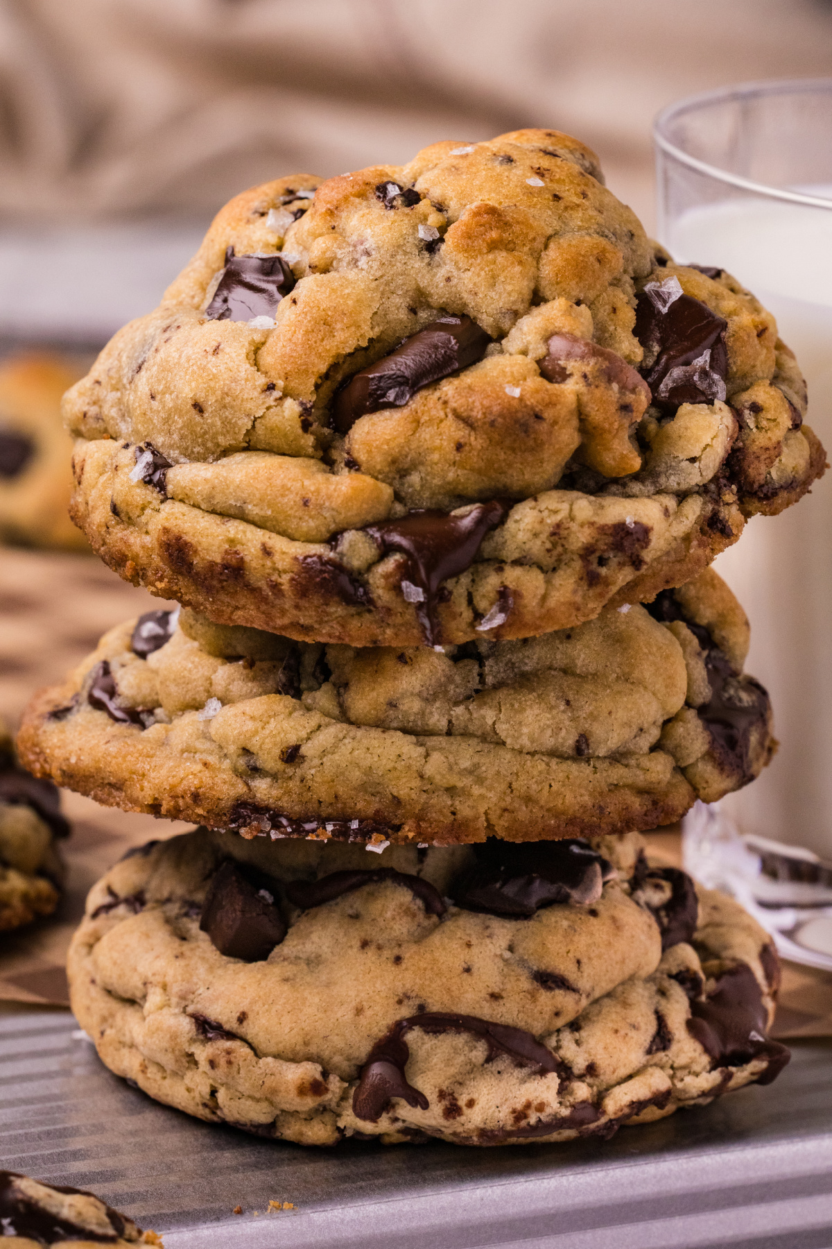 Three thick chocolate chip cookies stacked beside a glass of milk, with melted dark chocolate, golden tops, and flaky sea salt.