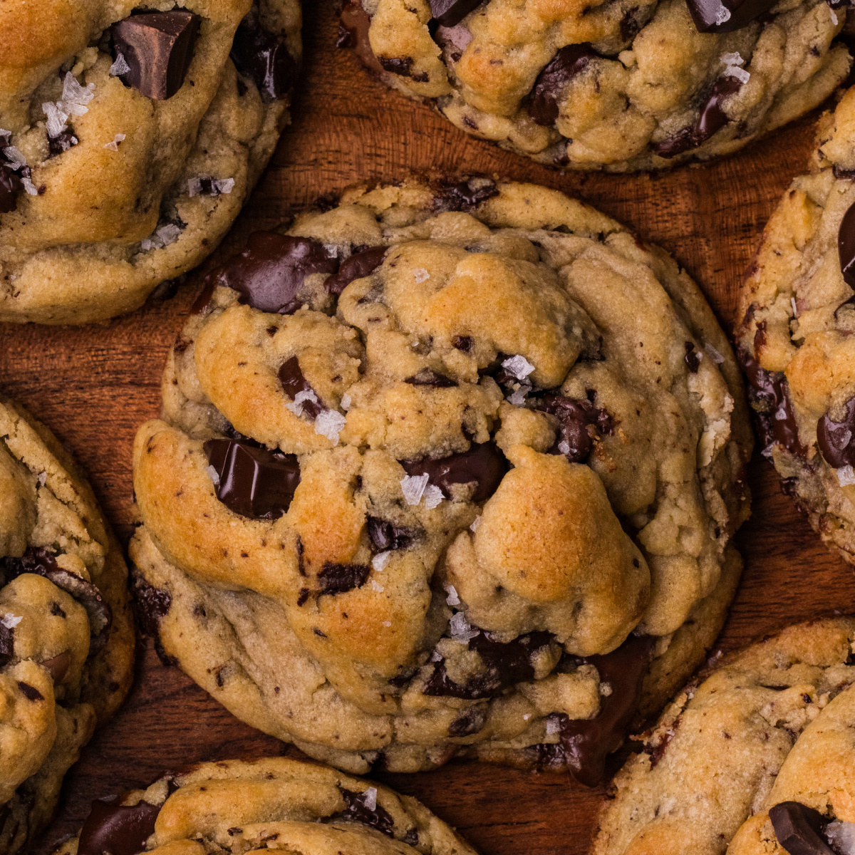 Close-up of a thick chocolate chip cookie on a wooden board with golden dough, dark chocolate chunks, and flaky sea salt.