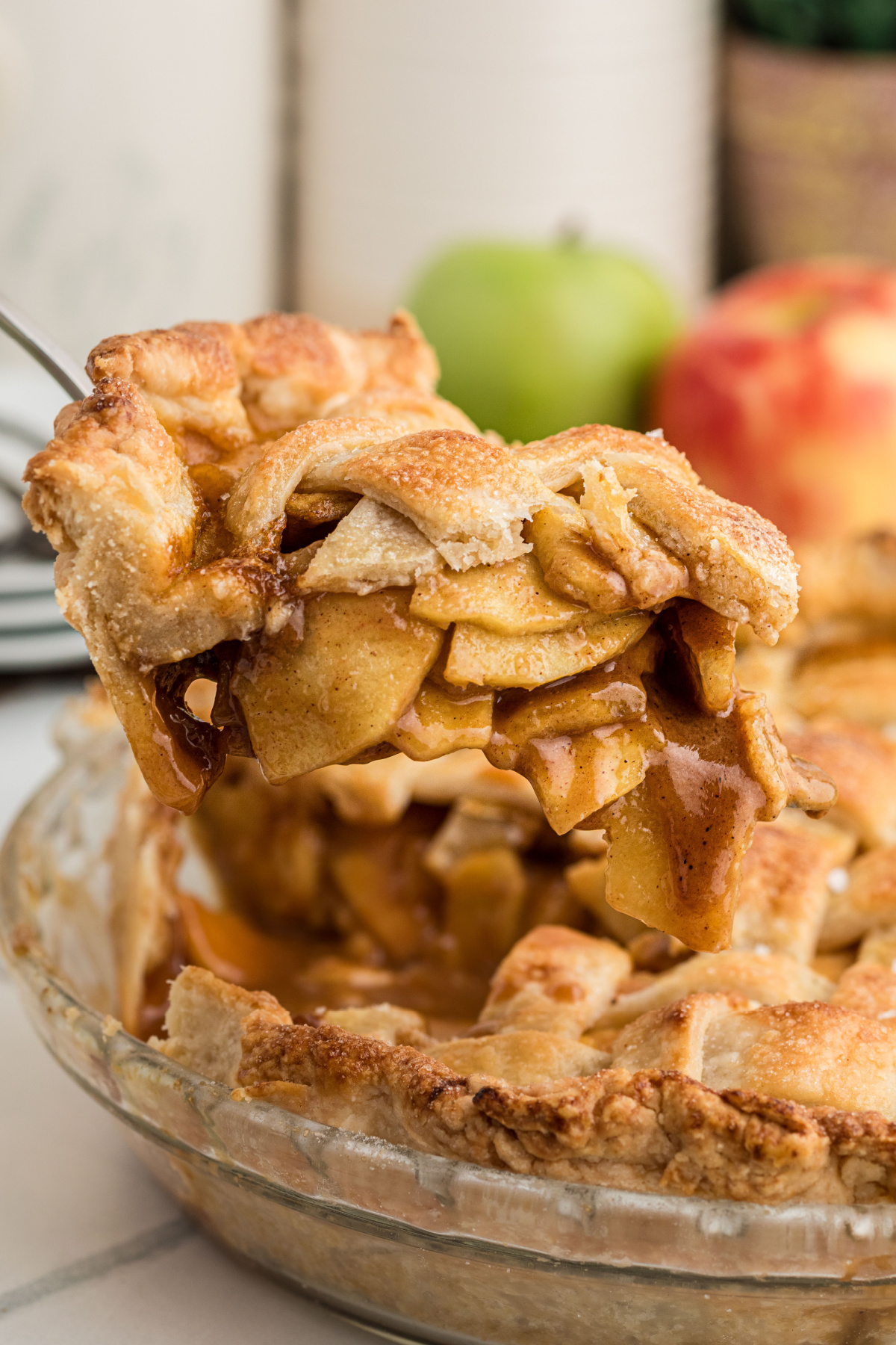 Slice of homemade apple pie lifted from a glass pie plate, showing thin apple slices coated in cinnamon-spiced caramel beneath a flaky lattice crust.