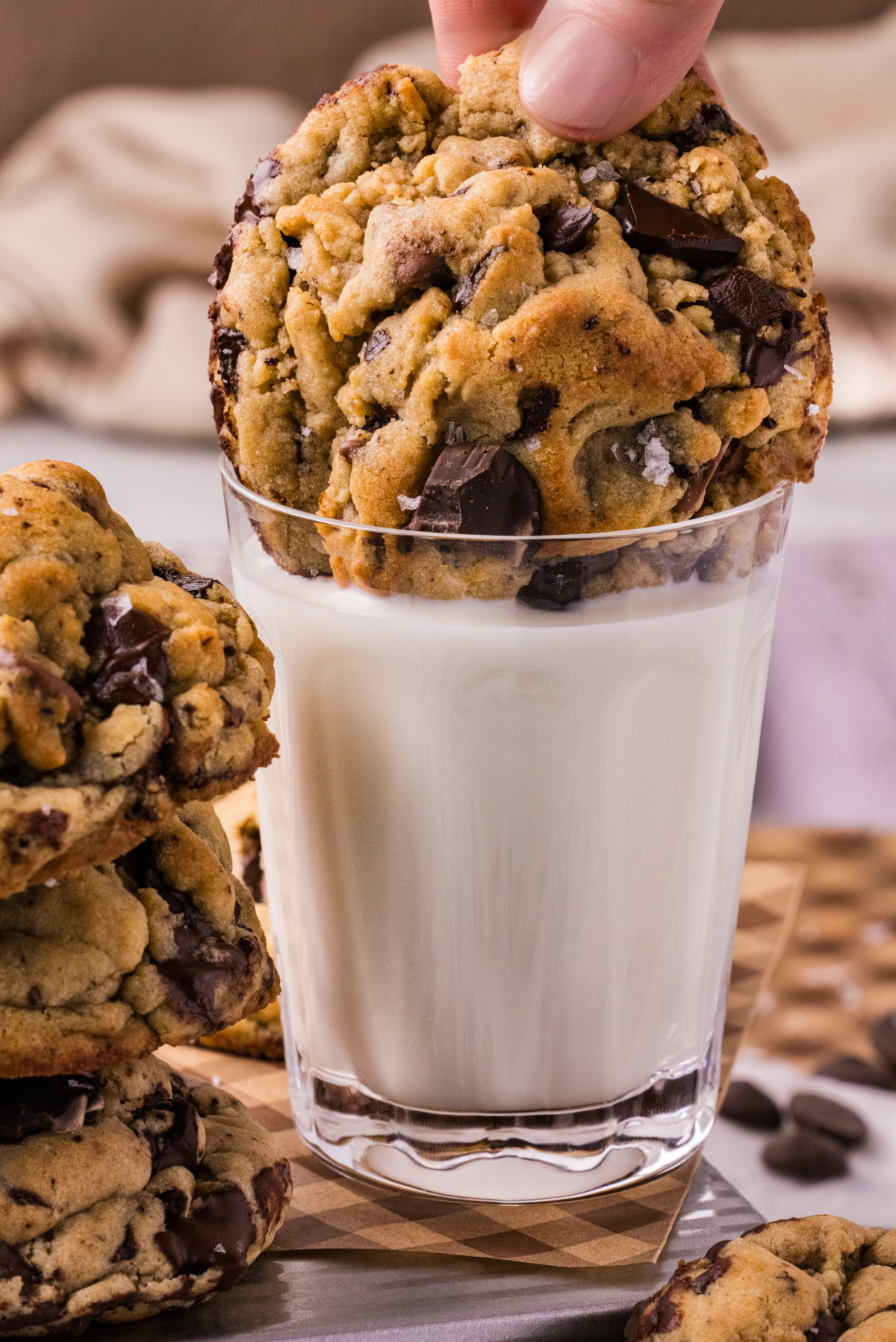 Hand dipping a thick chocolate chip cookie with dark chocolate chunks and flaky sea salt into a glass of milk.