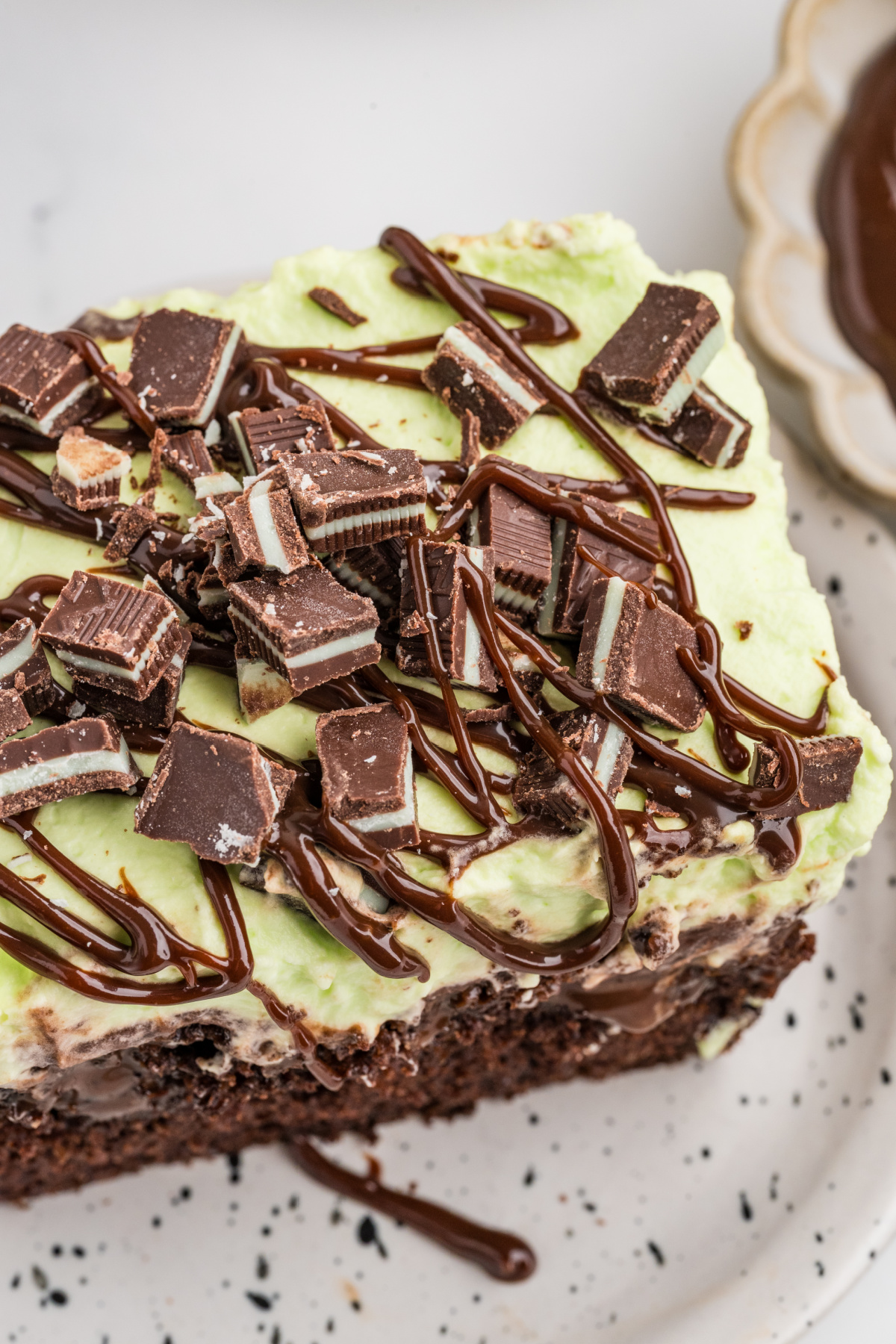 Close-up of chocolate poke cake topped with pale green mint whipped cream, chopped Andes mint chocolates and hot fudge drizzle.