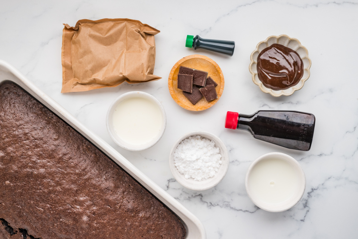 Ingredients for chocolate mint poke cake including baked chocolate cake, hot fudge sauce, pudding mix, milk, heavy cream, powdered sugar, mint extract, green food coloring and Andes mints.