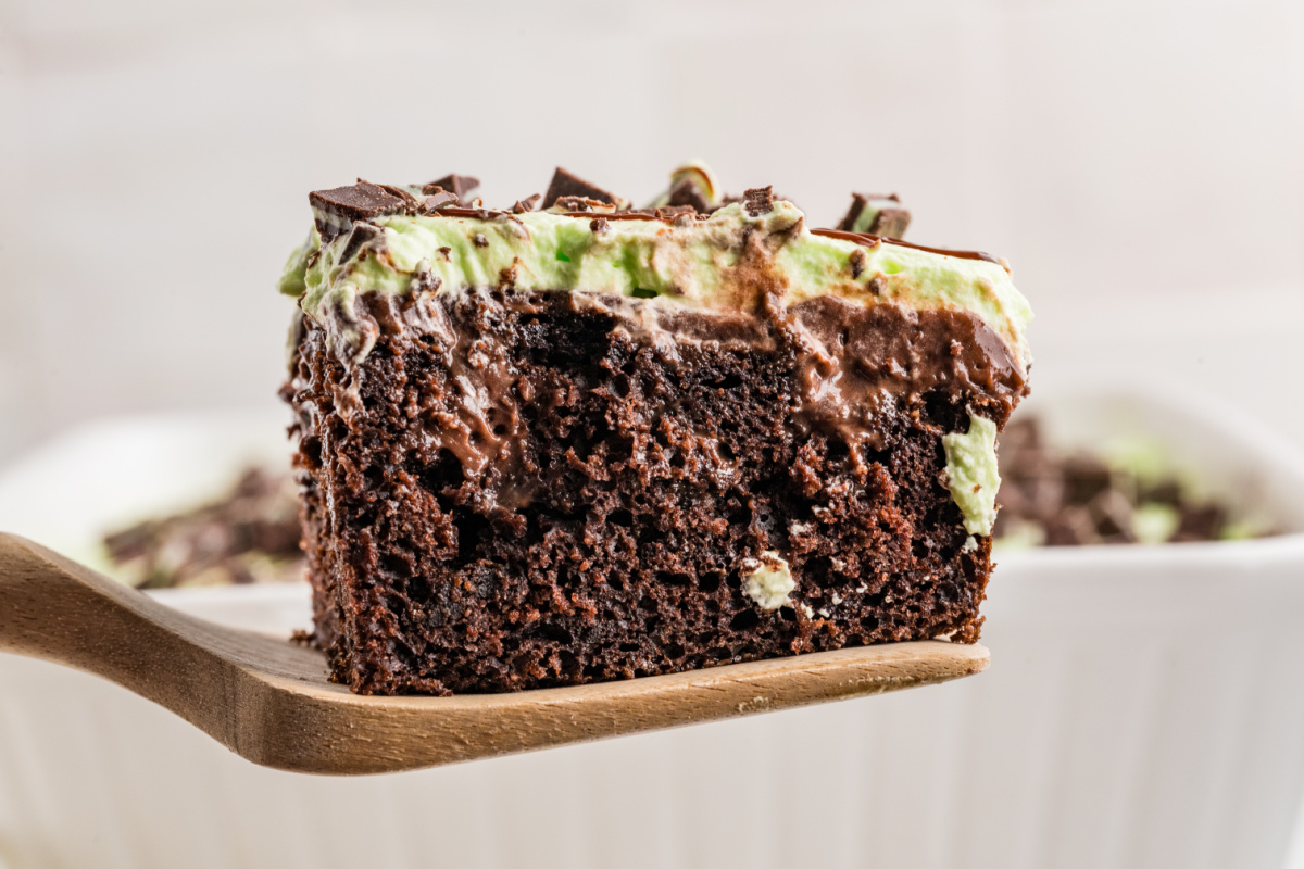 Slice of chocolate poke cake on a wooden spatula showing fudge and chocolate pudding inside, topped with mint whipped cream and Andes mints.