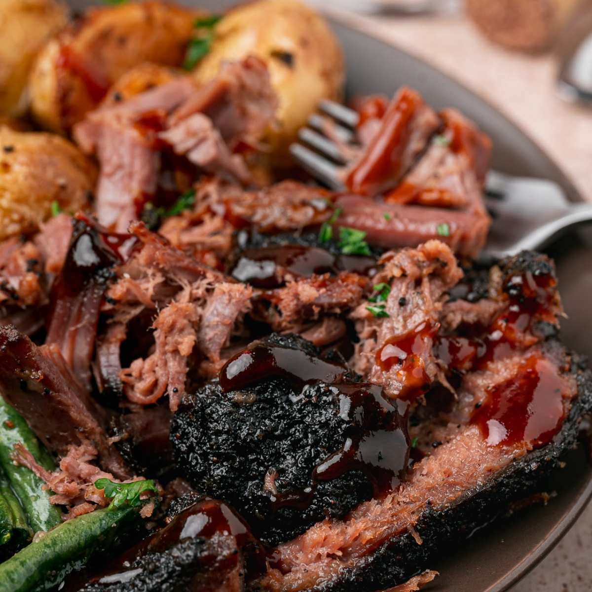 Coffee Rubbed Oven Brisket served with roasted baby potatoes, green beans, and barbecue sauce on a dark dinner plate.