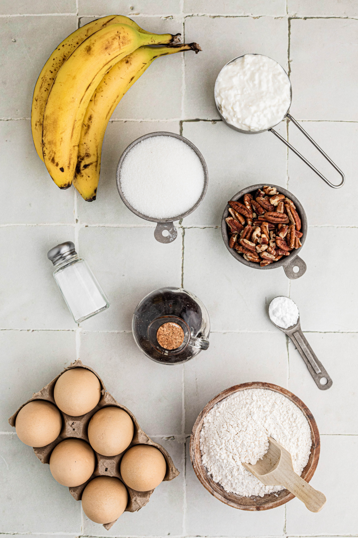 Ripe bananas, cottage cheese, flour, sugar, eggs, toasted pecans, vanilla, baking soda, and salt arranged on white tile.