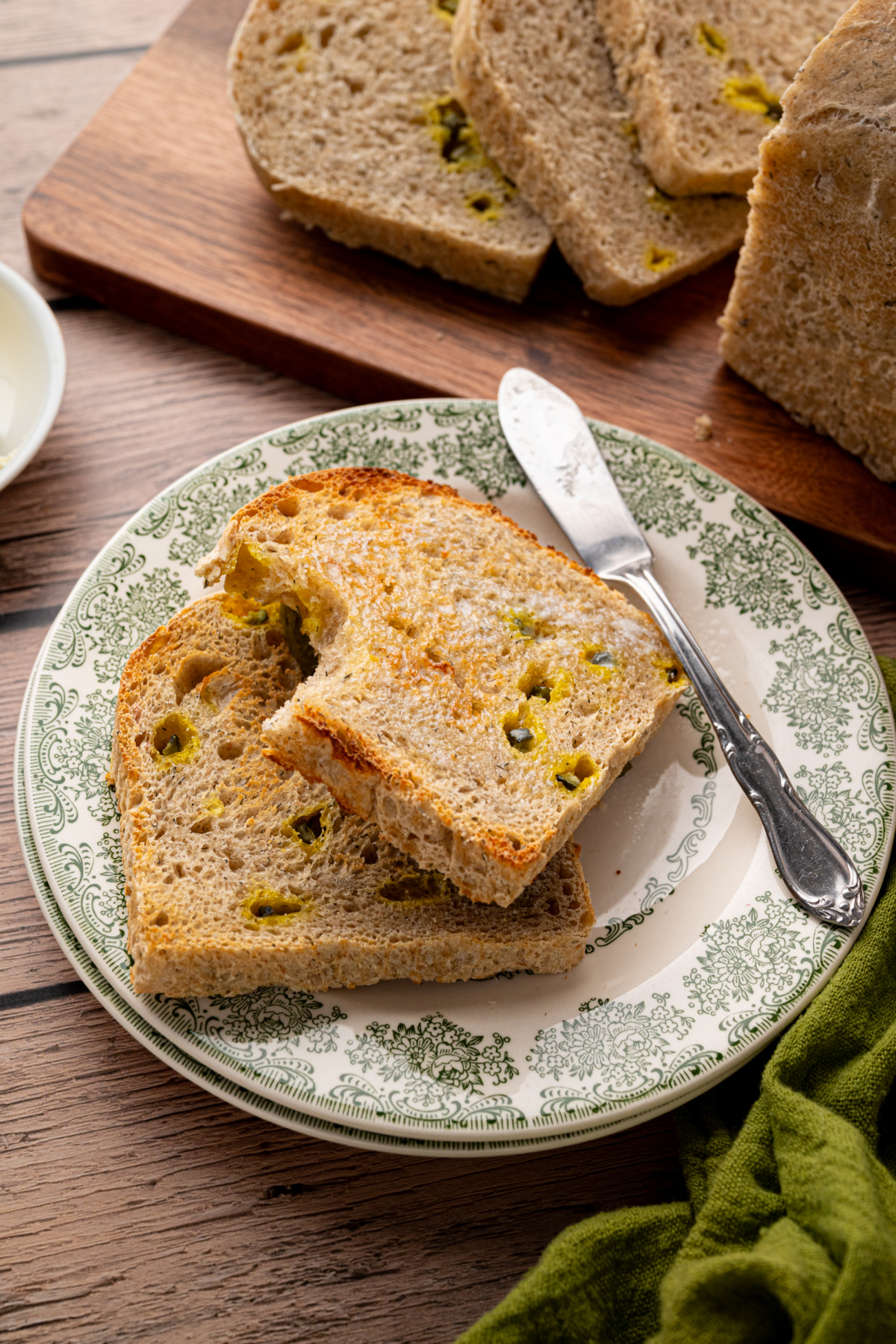 Two slices of toasted sourdough bread with chopped dill pickles and melted butter on a green patterned plate, with the sliced loaf in the background.
