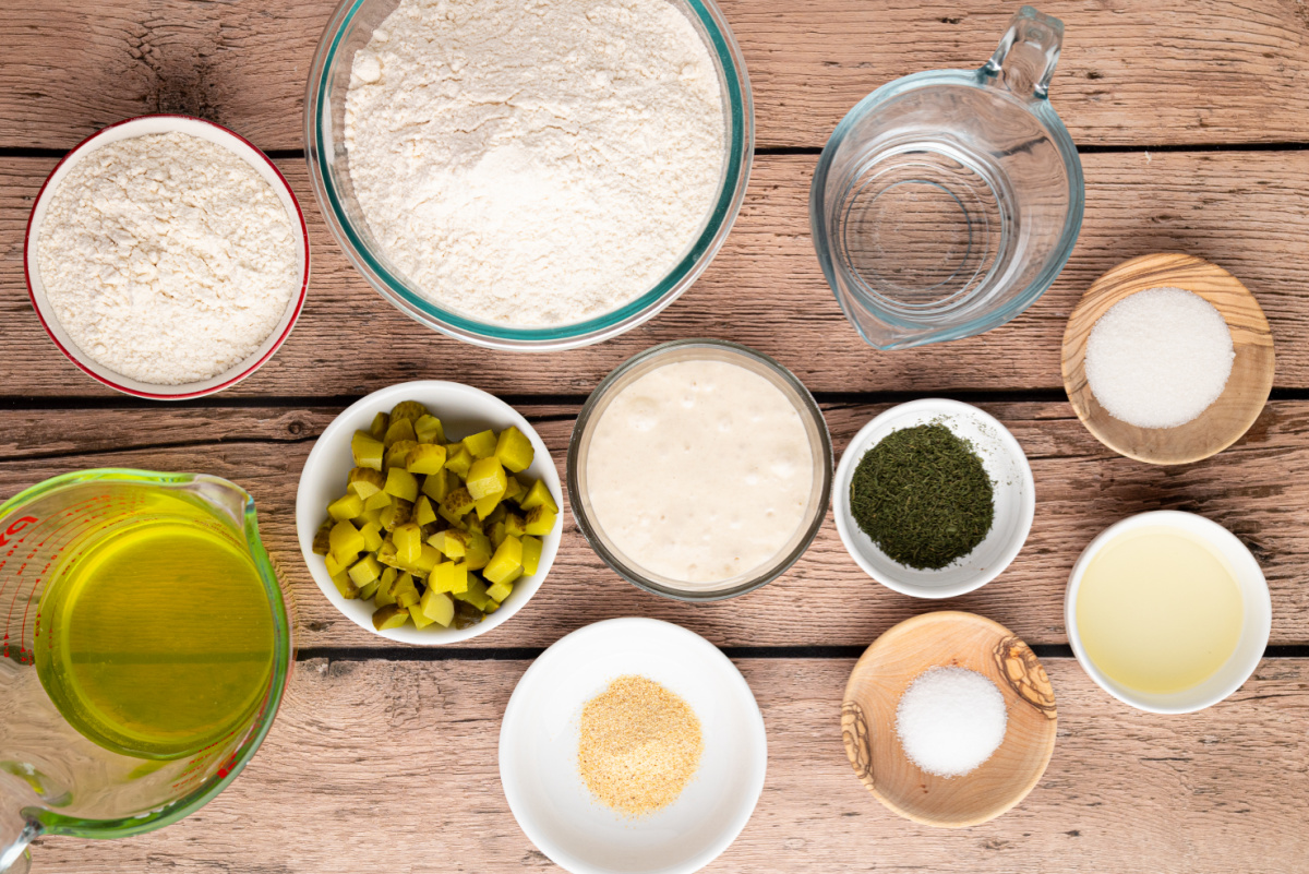 Ingredients for homemade sourdough pickle bread arranged on a wooden table, including chopped dill pickles, active starter, bread flour, whole wheat flour, pickle brine, dried dill, and garlic powder.