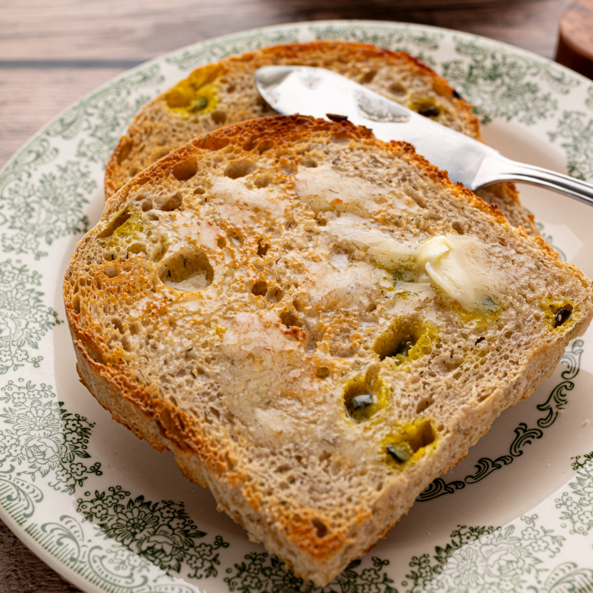 Close-up of toasted dill pickle sourdough bread spread with melting butter, showing chopped pickles and dried dill throughout the bread.