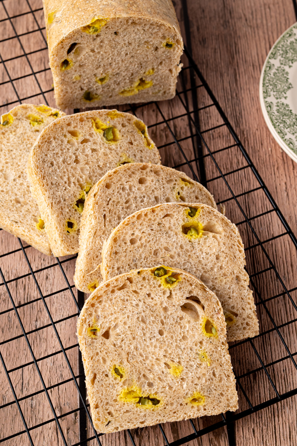 Sliced dill pickle sourdough sandwich bread arranged in a row on a cooling rack, showing the airy interior and pieces of chopped dill pickle.