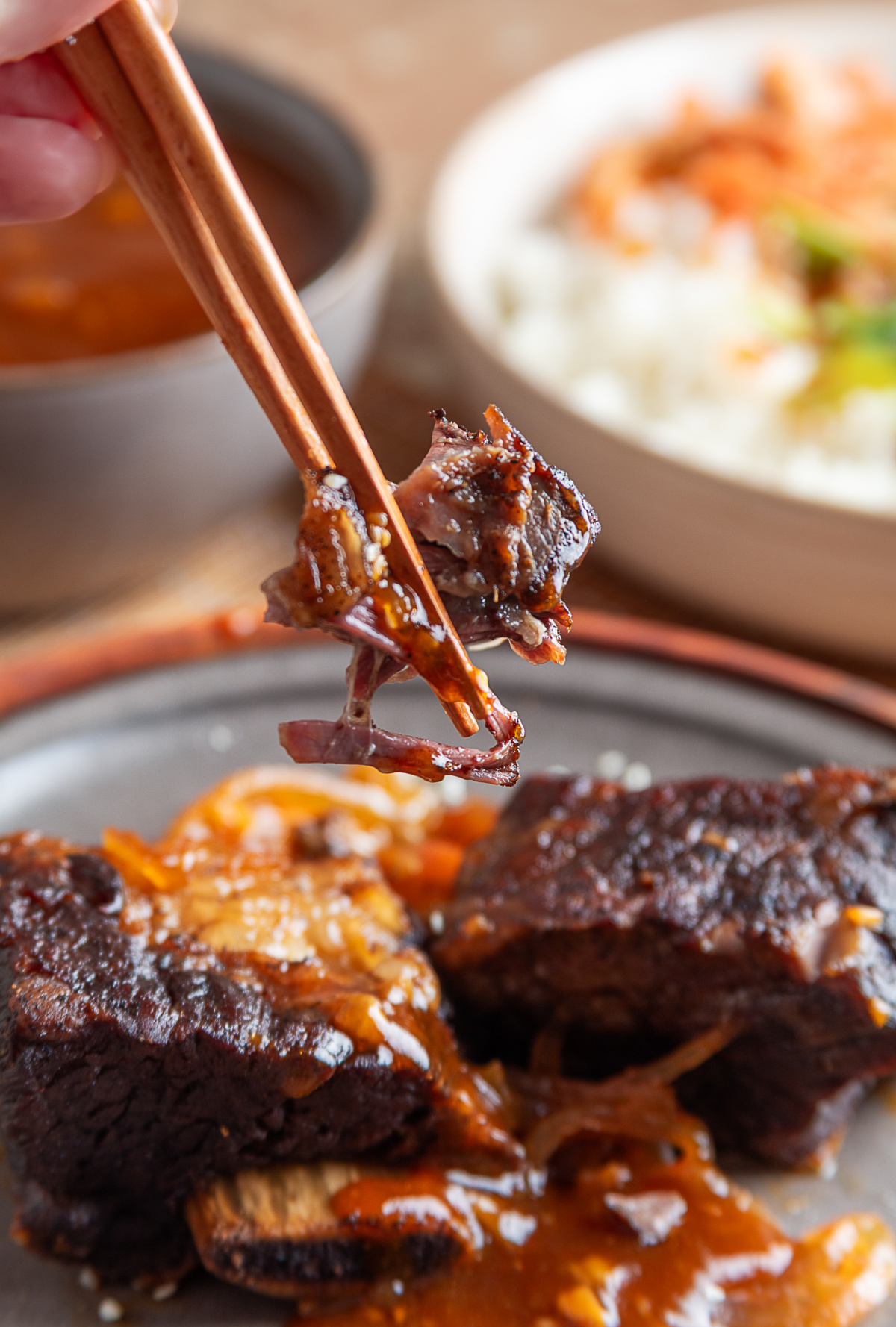 Chopsticks lifting a tender piece of slow-cooked beef short rib coated in gochujang sauce above the remaining ribs.