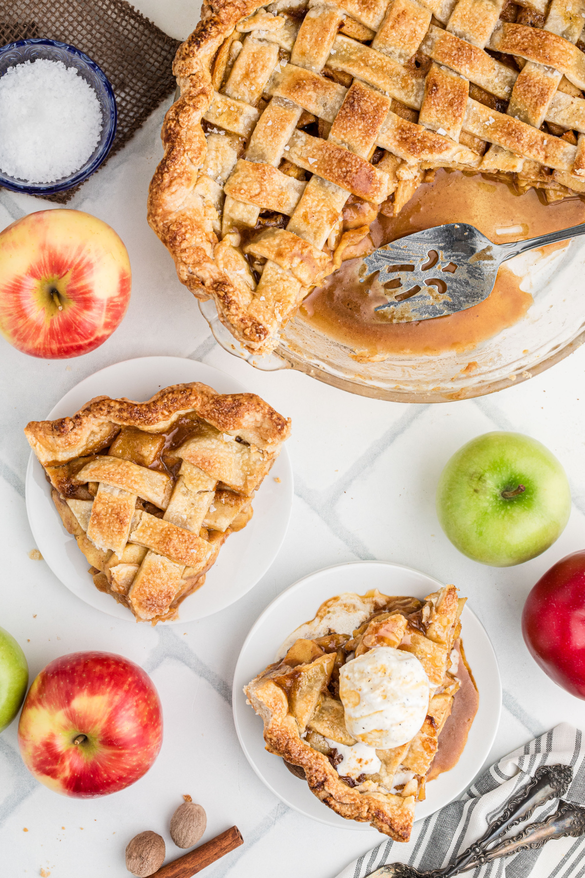 Overhead view of homemade caramel apple pie with a slice removed, two plated slices, vanilla ice cream, and caramel sauce pooled in the pie plate.