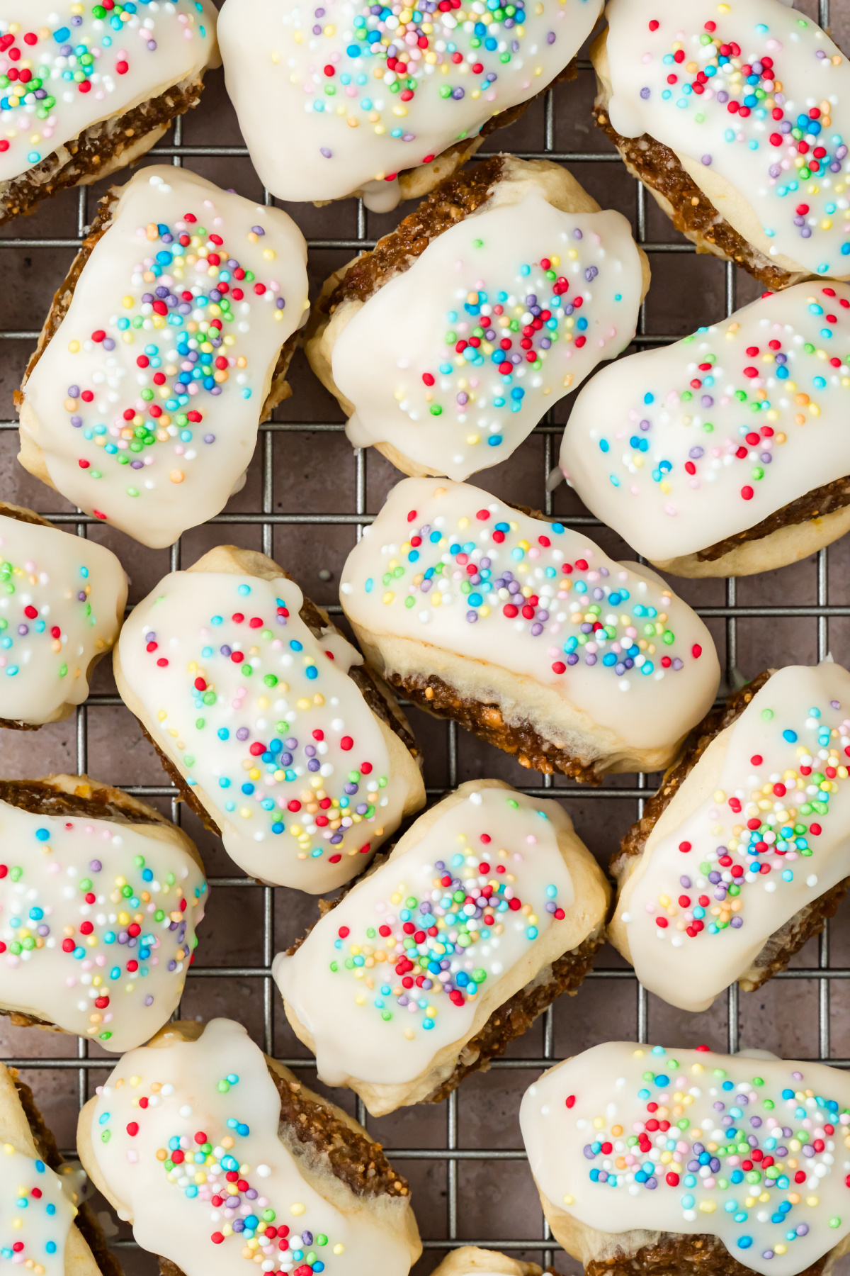 Italian Fig Cookies with Vanilla Icing and Colorful Nonpareils Overhead close-up of Italian fig cookies on a wire rack, showing thick white vanilla glaze, rainbow nonpareils, and fig filling along the sides.