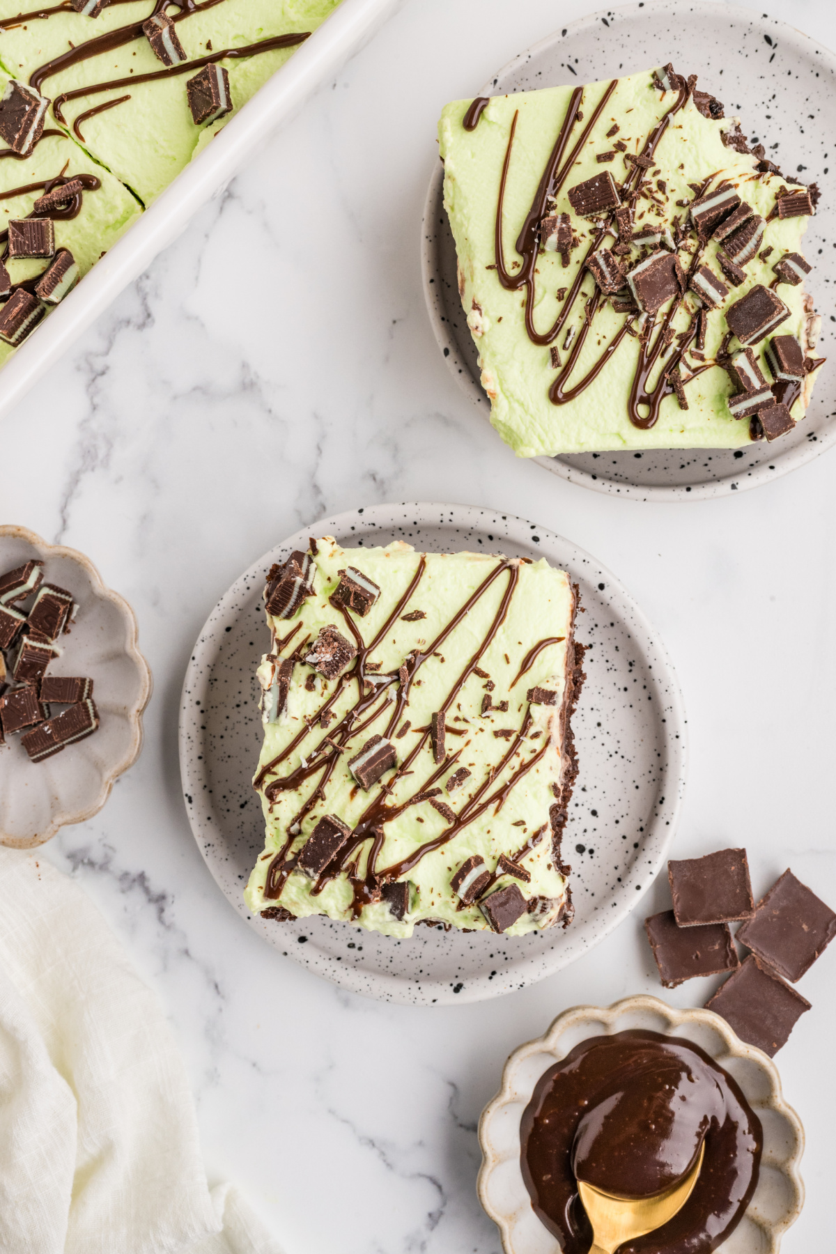 Two slices of mint chocolate poke cake on speckled plates with pale green whipped cream, chopped Andes mints and hot fudge drizzle.
