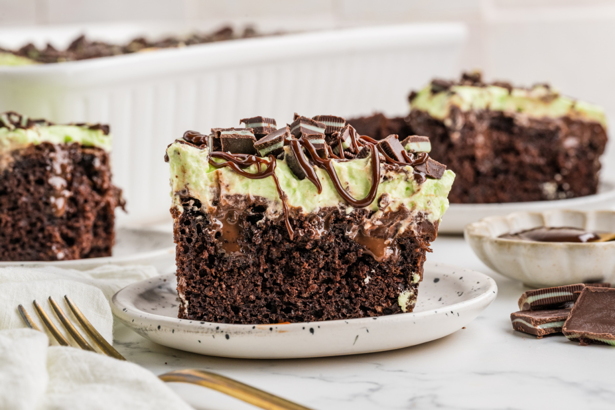 Slice of mint chocolate poke cake with hot fudge filling, green whipped cream, chopped Andes mints and chocolate drizzle.
