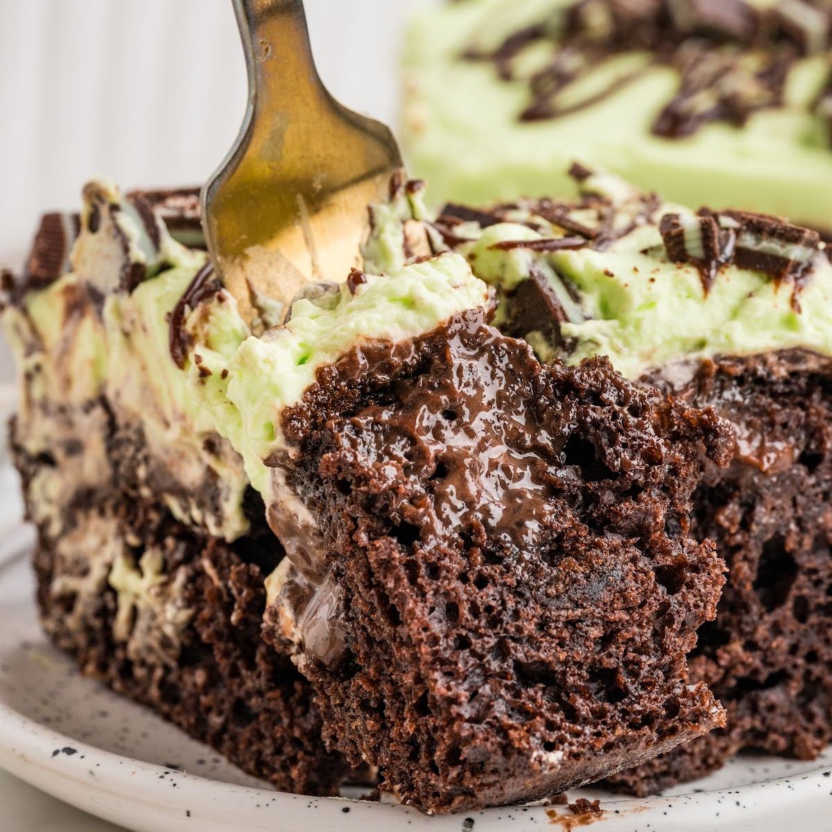 Close-up of mint chocolate poke cake with a fork lifting a piece of chocolate cake filled with hot fudge and topped with pale green mint whipped cream.