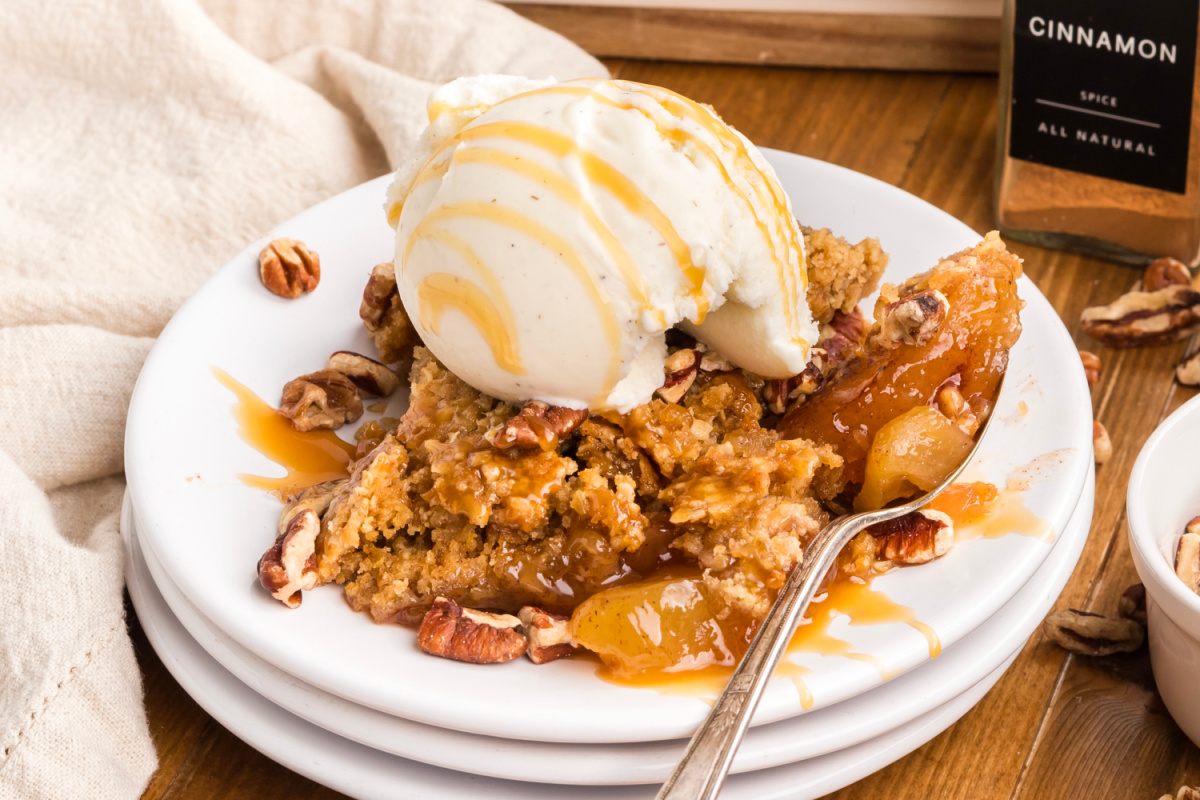 A serving of warm apple crisp with a golden oatmeal cookie crust, cinnamon apples, chopped pecans, caramel sauce, and vanilla ice cream. Oatmeal cookie apple crisp dump cake served with vanilla ice cream, caramel drizzle, chopped pecans, and cinnamon apple filling.