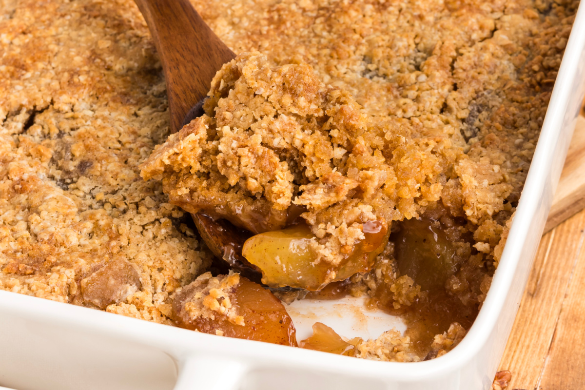 A serving of warm apple dump cake being lifted from the pan, revealing soft cinnamon apples beneath the golden oatmeal cookie topping. A spatula lifting oatmeal cookie apple crisp dump cake from a baking dish, showing the cracked cookie crust and cinnamon apple filling.