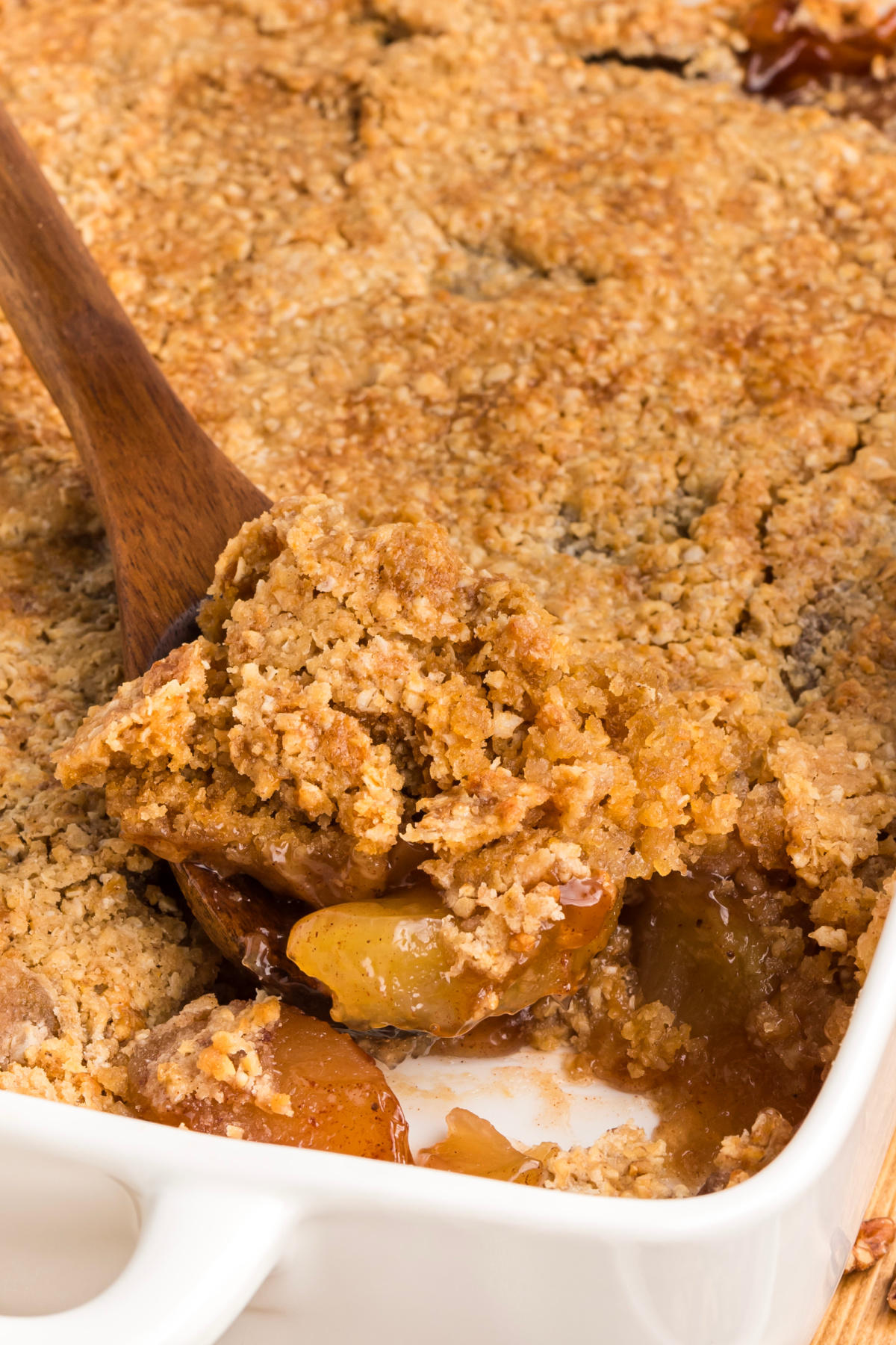 A close-up of the golden oatmeal cookie topping being lifted from the baking dish, revealing cinnamon-spiced apples underneath. Wooden spatula lifting a serving of apple crisp with a cracked oatmeal cookie crust and soft cinnamon apple filling.