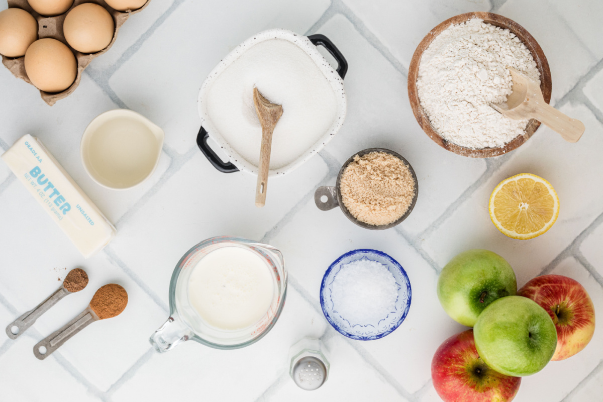 Ingredients for homemade apple pie arranged on a marble countertop, including Granny Smith and Honeycrisp apples, flour, butter, sugar, cream, lemon and spices.
