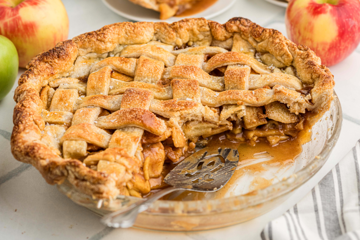 Salted caramel apple pie with a golden lattice crust and a slice removed, revealing thinly sliced apples and caramel sauce in a glass pie plate.