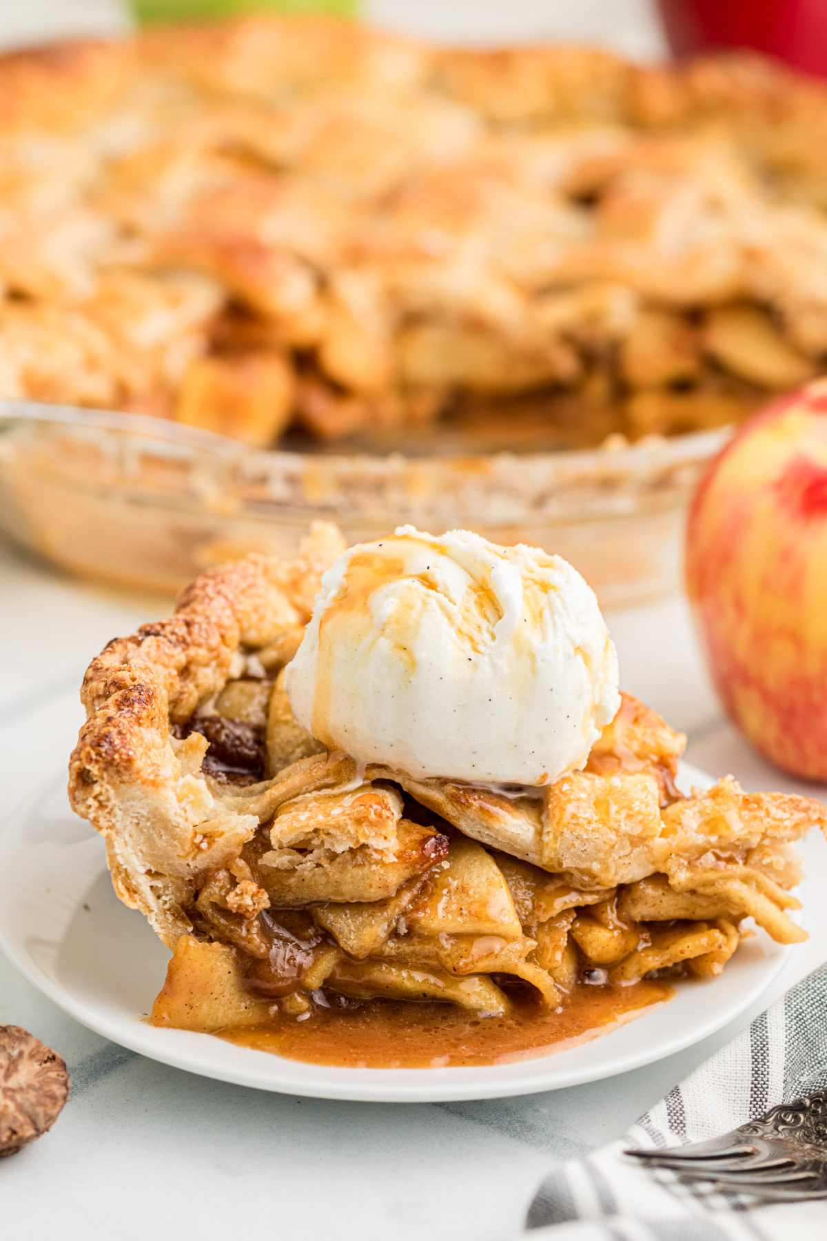 Slice of salted caramel apple pie with flaky lattice crust, thinly sliced apples, caramel sauce and a scoop of vanilla ice cream.