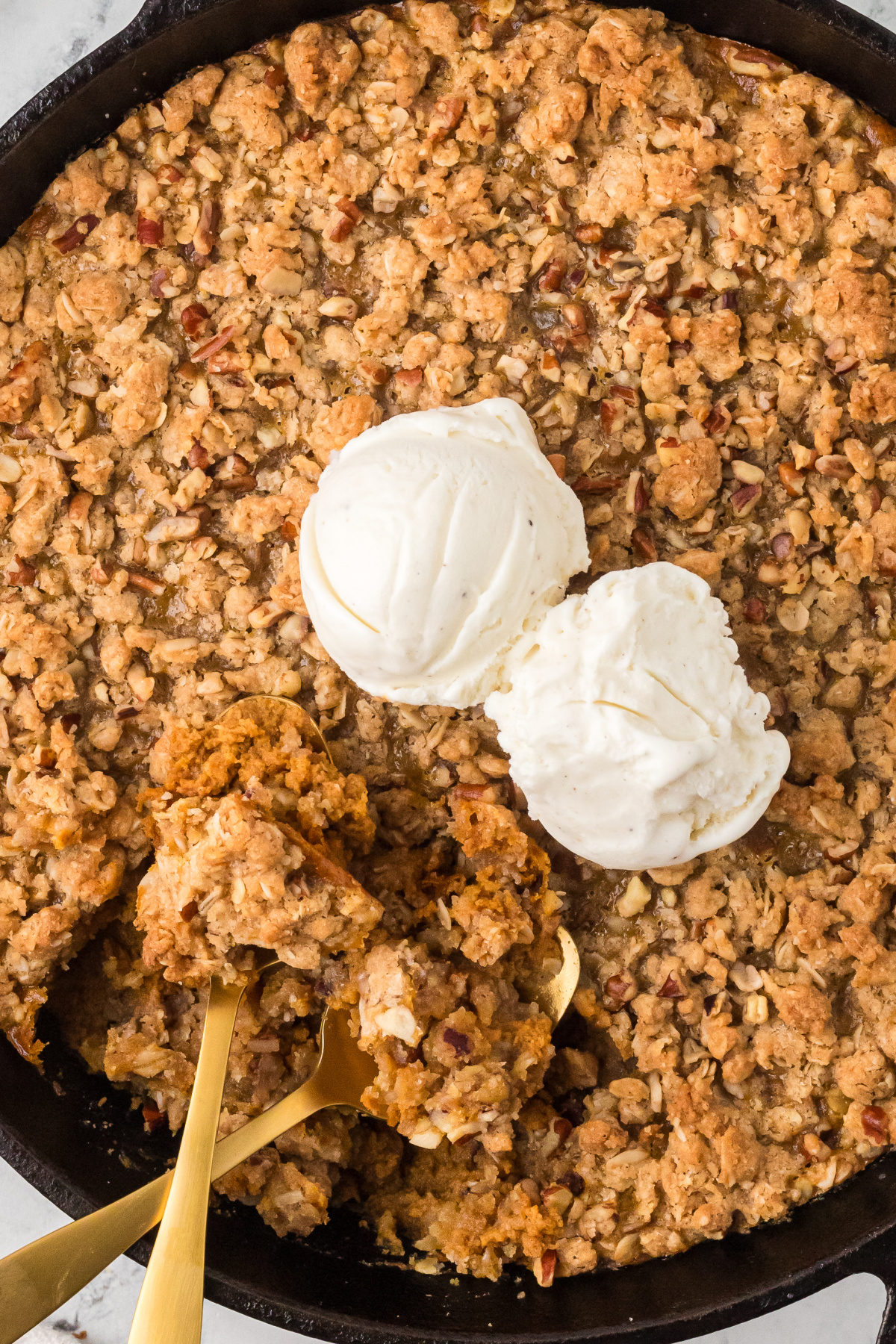 Warm pumpkin custard and buttery pecan oat topping served straight from the skillet with vanilla ice cream. Cast-iron skillet filled with pumpkin custard and oat pecan topping, topped with two scoops of vanilla ice cream and partially served with gold spoons.