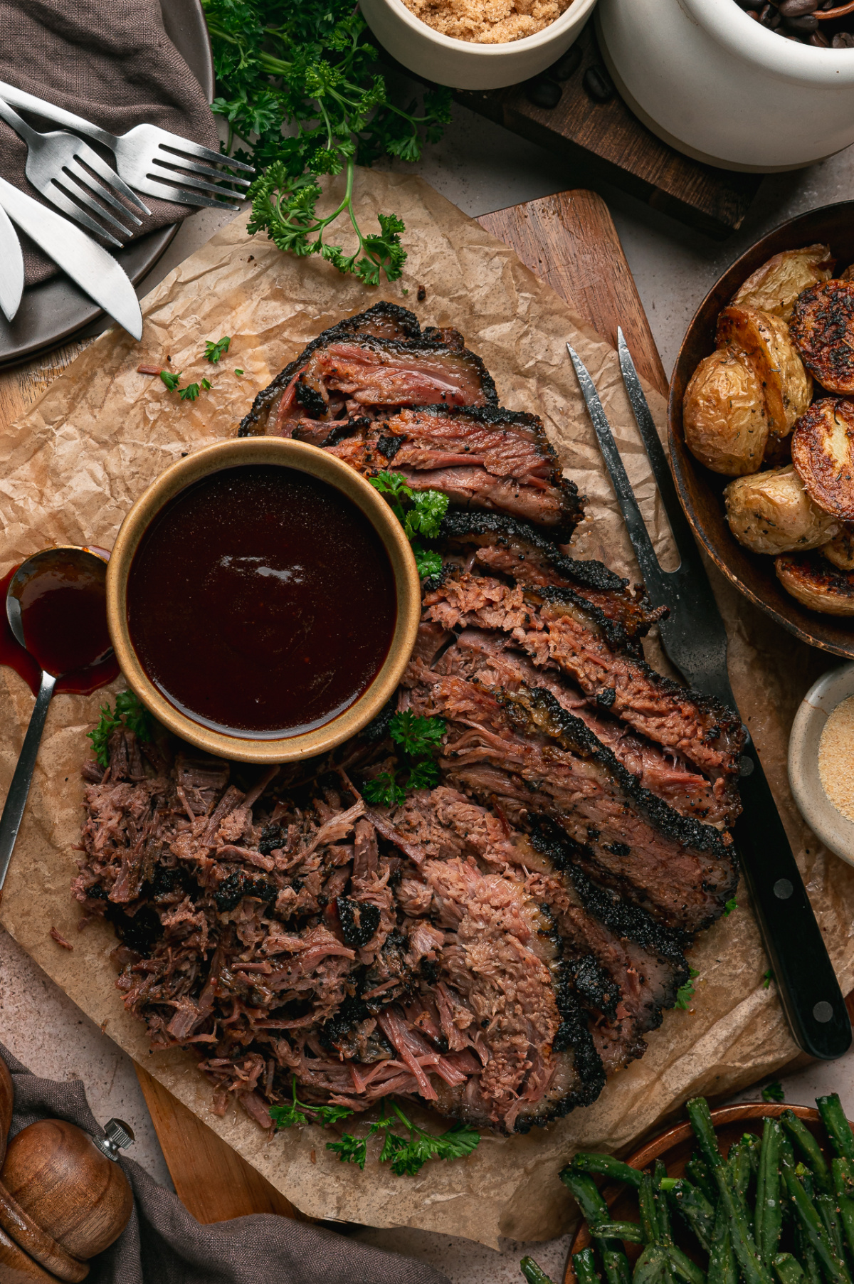Sliced and pulled Coffee Rubbed Oven Brisket arranged on a wooden serving board with barbecue sauce, roasted potatoes, and green beans.
