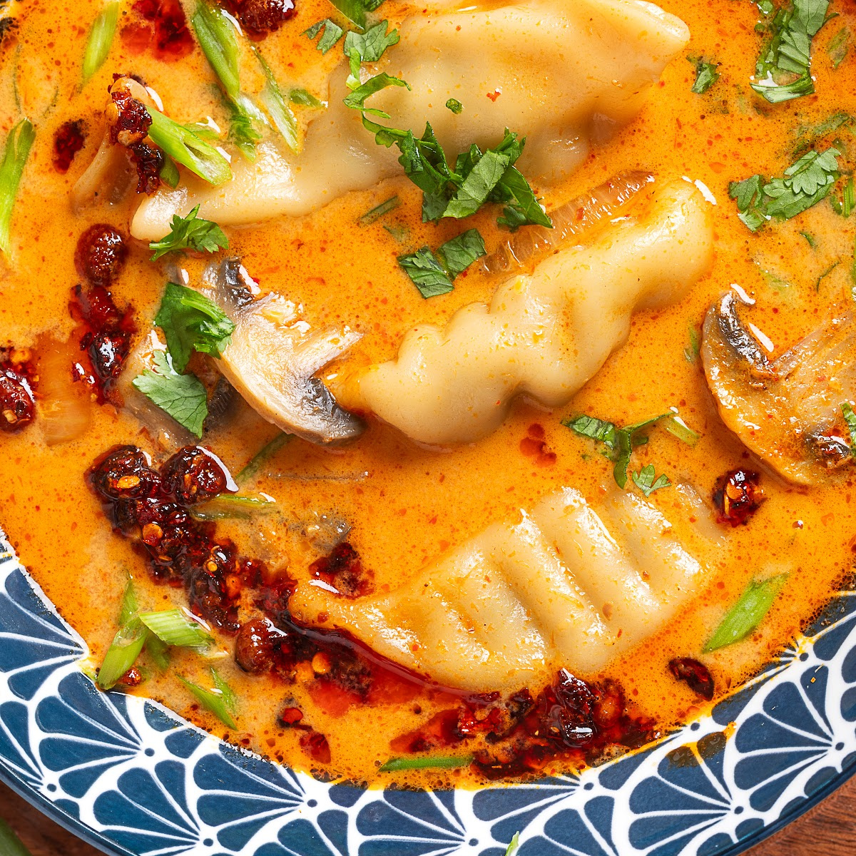 Thai Dumpling Soup with pork potstickers, sliced mushrooms, cilantro, and chili crisp in a creamy coconut red curry broth.
