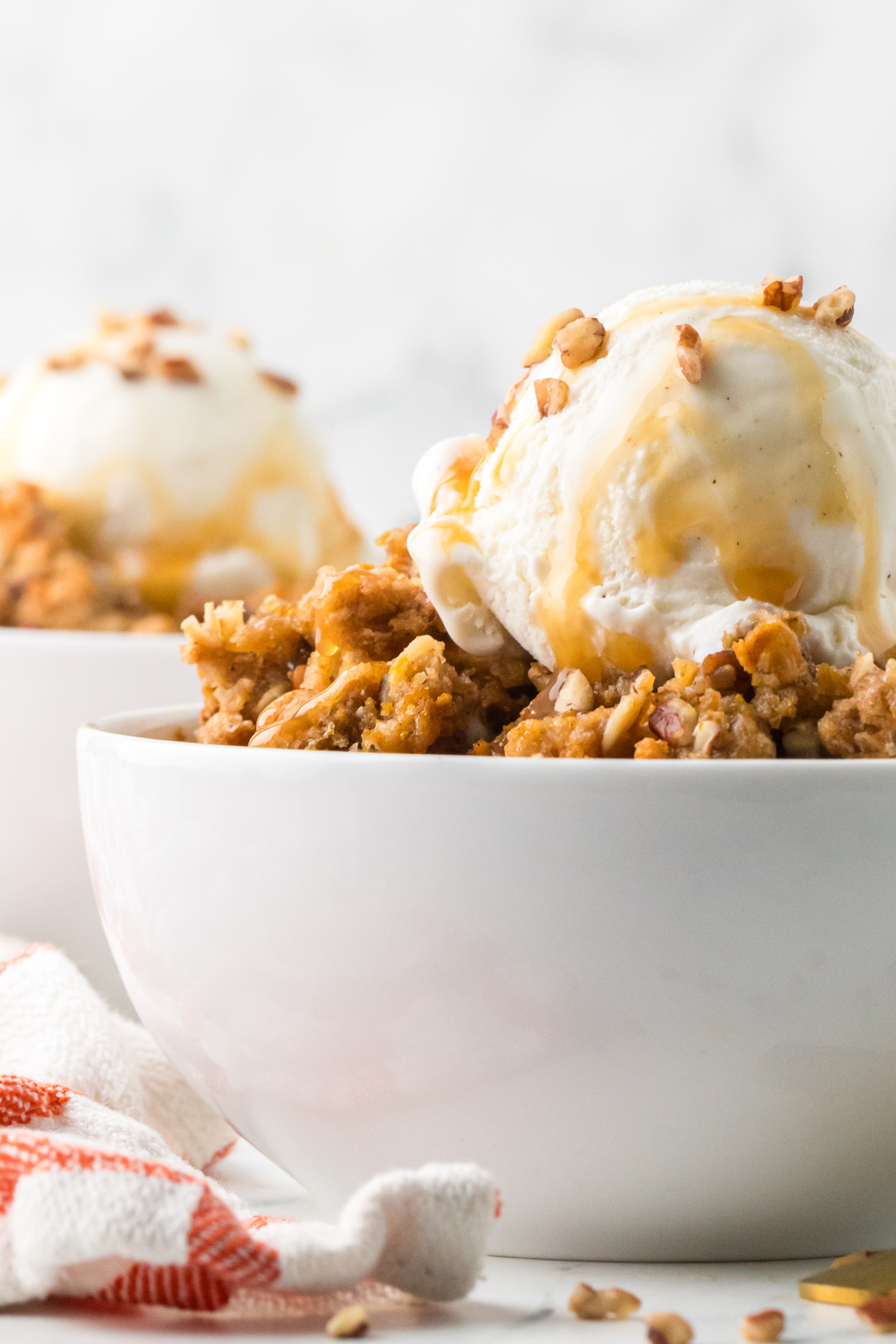 Vanilla ice cream melting over warm pumpkin custard with buttery oat clusters, chopped pecans, and caramel drizzle. Side view of a bowl of spiced pumpkin custard with oat and pecan topping, vanilla ice cream, and caramel sauce.