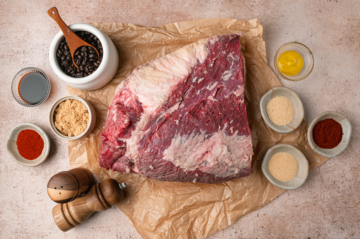 Raw whole packer beef brisket surrounded by ground coffee, brown sugar, mustard, liquid smoke, and dry rub seasonings.